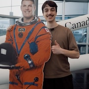 Le journaliste Tristan Champagne-Lessard pose aux c&ocirc;t&eacute;s d&rsquo;une r&eacute;plique en carton de l&rsquo;astronaute Jeremy Hansen.