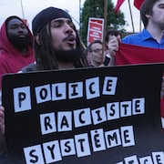 Dans une manifestation, un homme tient une affiche sur laquelle il est &eacute;crit &laquo; Police raciste syst&egrave;me complice &raquo;.