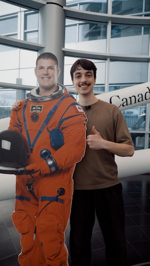 Le journaliste Tristan Champagne-Lessard pose aux côtés d’une réplique en carton de l’astronaute Jeremy Hansen.